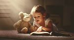 Symbolbild eines kleinen Mädchens mit geöffnetem Buch, das einem Teddybären vorliest. 