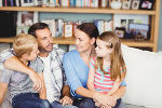 Symbolbild in Form einer Familie von zwei Elternteilen, die mit ihren Kindern auf der Couch sitzen und sich unterhalten.