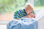 Ein Baby schläft, zugedeckt mit einer blauen Decke. Der größere Bruder (selbst noch ein Kleinkind), beugt sich darüber und gibt dem Baby einen Kuss. 