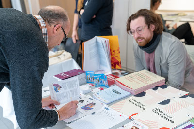Impressionen von den Marktständen, ein Mann notiert sich Informationen, vor ihm eine Menge an Broschüren Impressionen von den Marktständen, ein Mann notiert sich Informationen, vor ihm eine Menge an Broschüren