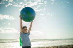Kind steht am Strand und hält einen Wasserball in der Form der Erdkugel in die Höhe.                                                                               