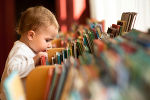 Ein Kleinkind steht vor einem Regal voller Bilderbücher und sieht sich diese an. 