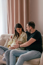 Ein paar sitzt auf einer Couch, die Frau ist schwanger und der Mann legt einen Arm um die Frau und den anderen auf den Babybauch.  