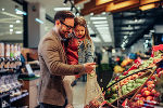 Einkaufender Vater mit der Tochter am Arm, das Obst wird in eine mitgebrachte Netz-Einkaufstasche gegeben. 